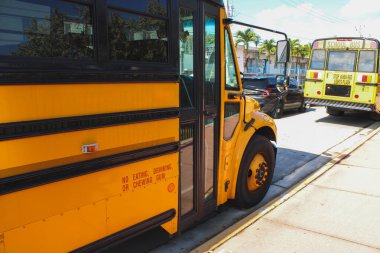Güneşli bir gökyüzünün altında şehir caddesi boyunca park etmiş parlak sarı bir okul otobüsünün yakın görüntüsü. Arka planda başka bir okul otobüsü görünüyor..