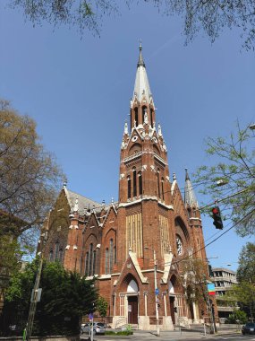 Çarpıcı tuğla cepheli ve keskin kulesi olan Gotik bir kilise açık mavi gökyüzüne karşı duruyor. Bina bir şehir kavşağına kurulmuş..