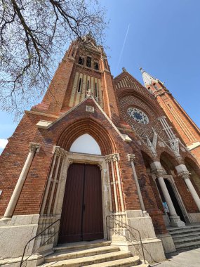 A low-angle view of a majestic brick Gothic-style church with tall spires, arched entrances, and intricate architectural details against a clear blue sky.