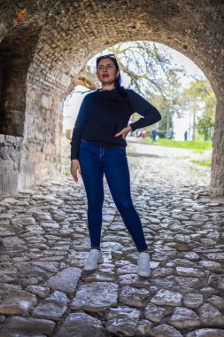 A confident young woman poses under an old stone archway on a cobblestone path. She wears a black sweater, blue jeans, and white sneakers, with trees and daylight visible behind the arch.