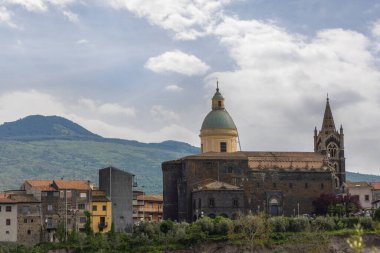 Yeşil kubbesi ve küçük bir İtalyan kasabasında Gotik çan kulesi olan tarihi kilise yeşil tepeler ve bulutlu gökyüzüne karşı kurulmuştur..