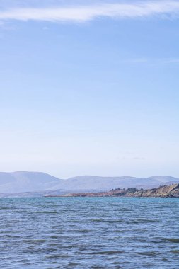 Mavi tonlarda deniz manzarası. Arka planda dağlar ve küçük bir ada görünüyor, dikey çekim