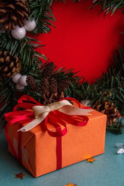 Noel çelenginin arka planında kırmızı fiyonklu turuncu ambalajlı Noel hediyesi.
