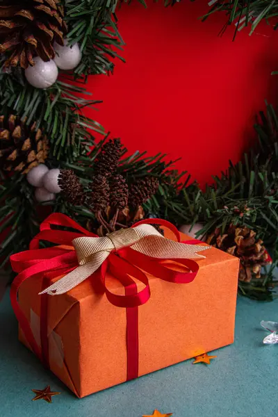 Noel çelenginin arka planında kırmızı fiyonklu turuncu ambalajlı Noel hediyesi.