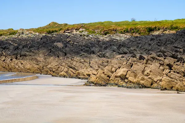 İnek ipi, Sherkin Adası, kayanın üzerinde koyu çizgili bir kayanın altındaki sahil, ön planda kum.