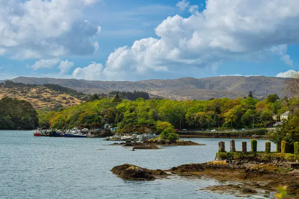 Sağda İrlanda 'nın Glengarriff kasabasındaki küçük bir körfezdeki Pierce manzarası, yosunlarla kaplı taş kalıntıları, arka planda ise arabalar ve dağların dış hatları var.