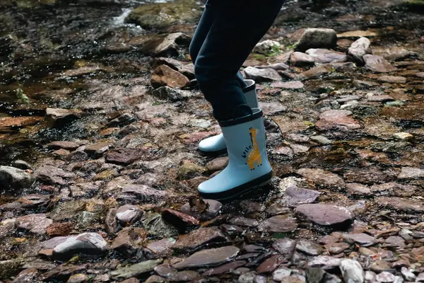 Nehrin taştan kıyısında lastik botlar içinde çocuk ayakları yürüyüş konsepti, nehir kenarında çocuk eğlencesi.