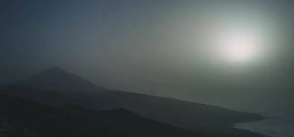 Karanlık bir manzara, arkasında bir dağ ve parlak bir güneş var. Görüntü sisle örtülmüş, gizemli bir atmosfer yaratıyor..