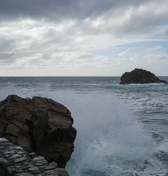 Dalgaların engebeli kayalara çarptığı dramatik bir deniz manzarası bulutlu bir gökyüzünün arka planına ve uzaktaki küçük bir adaya vuruyor..