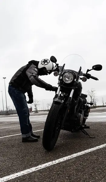 Koruyucu giysiler giymiş bir motosikletli, arka planda kasvetli bir atmosferle bir otoparkta sürüşe hazırlanırken görülüyor..