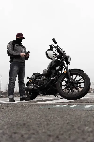 Ceket ve kot pantolon giyen bir adam telefonunu kontrol ederken siyah bir motosikletin yanında duruyor. Sahne bir duraksama ya da bekleme anını yakalar.