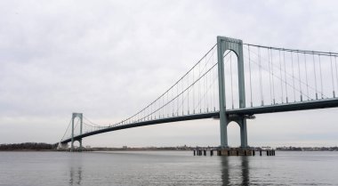 Görüntü, bulutlu bir günde bir su kütlesinin üzerine uzanan asma köprü tasvir ediyor. Köprünün kabloları ve destekleri belirgin bir şekilde görülüyor.