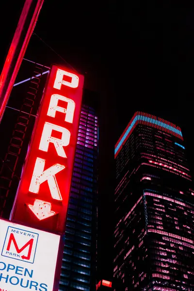 Görüntü bir park levhası ve geceleri aydınlık pencereli bir gökdelen gösteriyor. Sahne şehir yaşamını ve mimarisini yakalıyor..