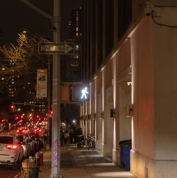 Tek yönlü bir tabela ve yaya geçidi sinyali olan bir şehir caddesi sahnesi. Cadde binalarla ve arabalarla dolu. Yolun kenarına park edilmiş..