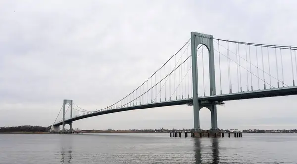 Görüntü, bulutlu bir günde bir su kütlesinin üzerine uzanan asma köprü tasvir ediyor. Köprünün kabloları ve destekleri belirgin bir şekilde görülüyor.