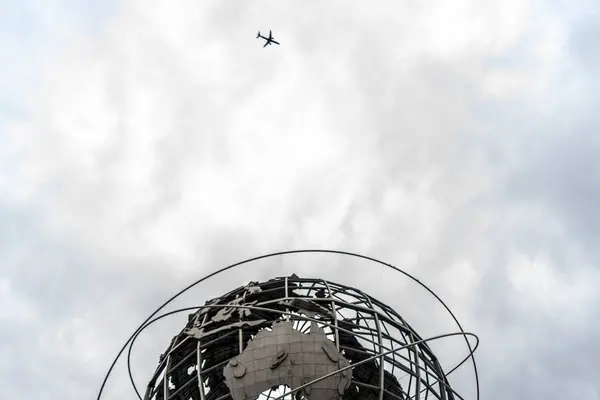 Görüntü, bulutlu bir gökyüzü ve uzaktan uçan bir uçağın zeminine dayanan karmaşık bir iskelete sahip büyük bir metal küre heykelini içeriyor. Heykel önemli bir sanat eseri veya simgesi gibi görünüyor..