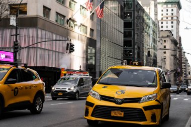 Kalabalık bir caddede diğer araçlar ve arka planda yüksek binalarla birlikte sarı bir taksi sürüyor. Sahne, şehir hayatının koşuşturmasını yakalıyor..