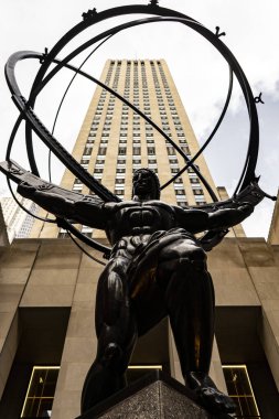 Prometheus 'un geniş bir heykeli, uzun bir gökdelenin önünde duran bir küreyi tutan kolları açık bir heykeli. Heykel koyu metalden yapılmış gibi görünüyor ve bir kaideye yerleştirilmiş..