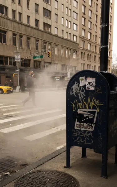 Grafiti ile kaplı bir posta kutusu şehir ortamında bir kaldırımda yer almaktadır. Arka planda karşıdan karşıya geçen biri var..