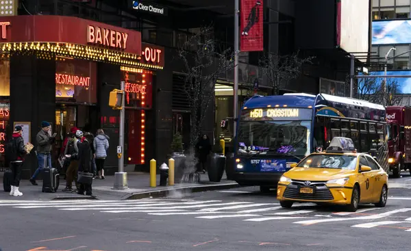 Sarı taksisi ve mavi otobüsü olan hareketli bir şehir caddesi kavşaktan geçiyor, bir fırın ve restoranın önündeki kaldırımda yürüyen insanlarla çevrili..