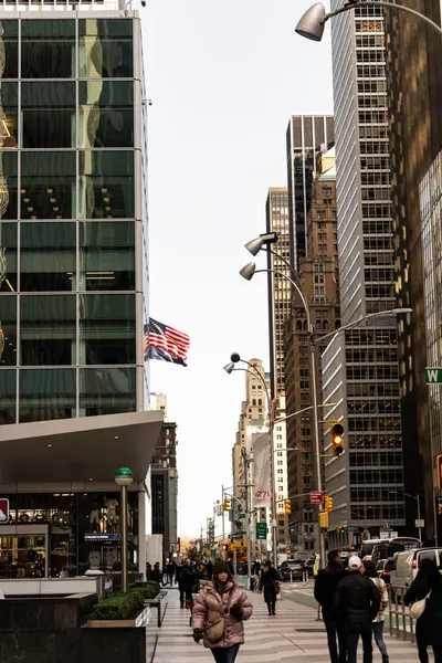 Uzun binaların ve kaldırımda yürüyen insanların olduğu bir şehir caddesi sahnesi. Arka planda Amerikan bayrağı görünüyor..