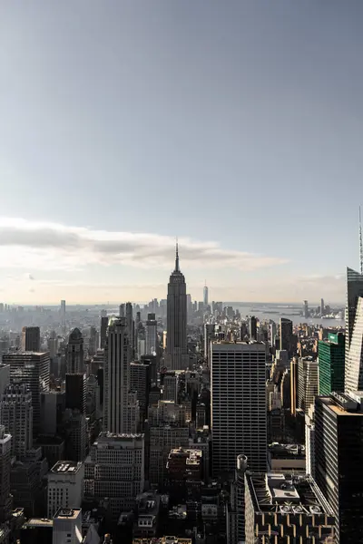New York şehrinin hareketli ufuk çizgisinin havadan görünüşü ve ikonik Empire State Binası dimdik ayakta duruyor. Şehrin yoğun gökdelenleri parçalı bulutlu bir gökyüzünün altında ufka doğru uzanıyor..