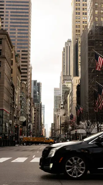 Uzun gökdelenler ve Amerikan bayraklarıyla dolu, önünde siyah bir araba ve arkasında sarı bir taksi olan bir şehir caddesi..