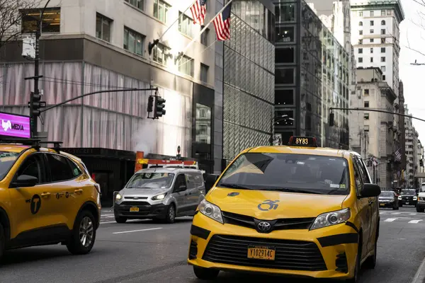 Kalabalık bir caddede diğer araçlar ve arka planda yüksek binalarla birlikte sarı bir taksi sürüyor. Sahne, şehir hayatının koşuşturmasını yakalıyor..