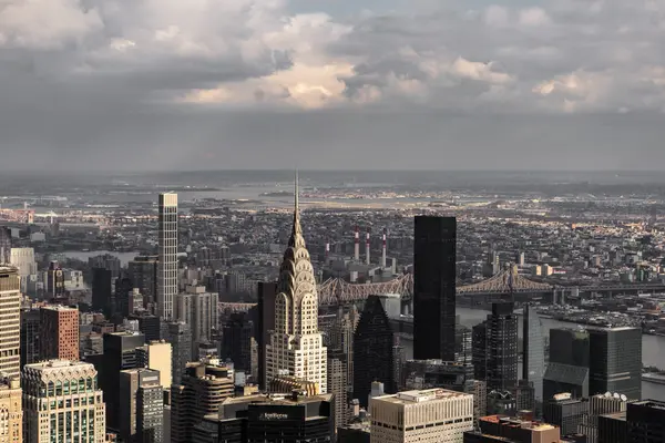 Bu görüntü, ikonik gökdelenler ve bulutlu bir gökyüzünün yer aldığı New York şehrinin ufuk çizgisinin çarpıcı hava görüntüsünü yakalıyor. Şehrin yoğun mimarisi ve görkemli binaları sergileniyor..