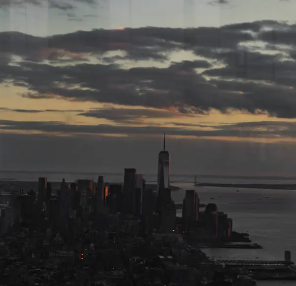 New York şehrinin karanlık bir manzarası. Güneş bulutların ardında batıyor. Yükselen gökdelenlere ve rıhtıma sıcak bir parıltı saçıyor..