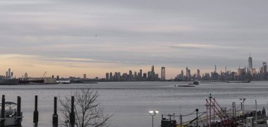 Önünde tekne ve rıhtım bulunan bir su kütlesi üzerinde bir şehrin ufuk çizgisi görülüyor. Sahne, bulutlu bir gökyüzüne karşı şehrin siluetiyle bir sükunet anı yakalıyor..