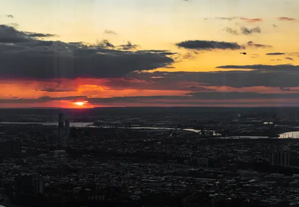 Güneş bulutlu bir gökyüzünün ardında batarken şehir manzarasının nefes kesici bir görüntüsünü yakalıyor. Şehir manzarasına sıcak turuncu bir ışık saçıyor. Şehrin silueti, gökyüzünün canlı renkleriyle çelişiyor..