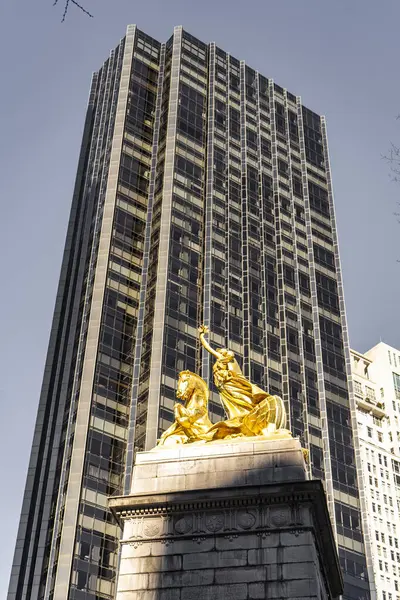 Görüntü, yansıtıcı pencereleri olan yüksek bir gökdelenin önünde duran taş bir kaidede sersemletici altın bir heykeli gösteriyor. Antik heykel ve modern bina arasındaki zıtlık çarpıcı bir görsel etki yaratır..