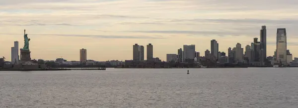 New York City siluetinin panoramik görüntüsü solunda duran ikonik Özgürlük Heykeli. Şehrin yükselen gökdelenleri ufuk boyunca uzanır, bulutlu bir gökyüzüne karşı kurulur..