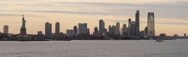 İkonik Özgürlük Heykeli, sakin bir günbatımında Manhattan 'ın görkemli siluetine bakan solda dimdik ayakta duruyor. Ön plandaki sakin sular sahnenin sükunetini artırıyor..