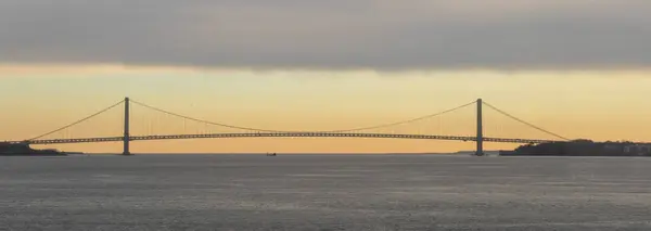 Görüntü, gün batımında bir su kütlesi boyunca uzanan asma köprünün sakin bir görüntüsünü yakalıyor. Köprünün silueti gökyüzünün canlı renklerine karşı duruyor..