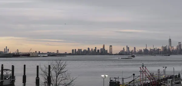 Önünde tekne ve rıhtım bulunan bir su kütlesi üzerinde bir şehrin ufuk çizgisi görülüyor. Sahne, bulutlu bir gökyüzüne karşı şehrin siluetiyle bir sükunet anı yakalıyor..