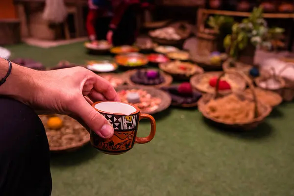 Görüntü, yeşil bir yüzeyde farklı yiyeceklerle dolu tabaklarla çevrili, karmaşık bir tasarımı olan canlı bir çay bardağını tutan kişiyi gösteriyor..