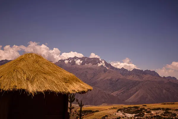 Sazdan bir kulübe, engebeli dağların çarpıcı zeminine ve açık mavi gökyüzüne karşı, önplanda huzur içinde duruyor. Huzurlu sahne, doğayla bir uyum ve sükunet hissi yayar..