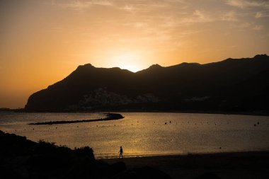 Gün batımında plajda yürüyen bir kişinin sakin bir sahnesi. Arka planda görkemli bir dağ sırası ve batan güneşin ılık parıltısı suya altın bir ışık saçıyor..