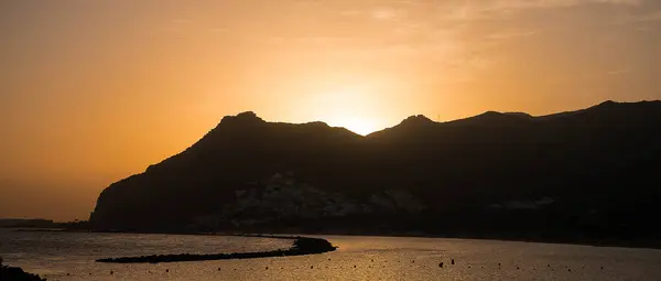 Güneş bir dağ sırasının arkasında batıyor. Önünde küçük bir ada olan huzurlu bir su kütlesinin üzerine ılık bir parıltı saçıyor..