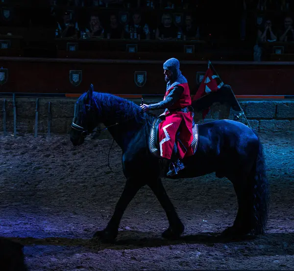 Kırmızı zırhlı bir ortaçağ şövalyesi loş bir arenada siyah bir ata biniyor.