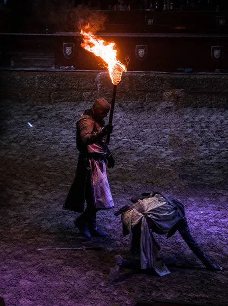 Başka biri karanlık bir arenada yerde yatarken pelerinli bir kişi yanan bir meşale tutar. Sahnenin ortaçağ atmosferi var, dramatik ya da yoğun bir anı çağrıştırıyor..