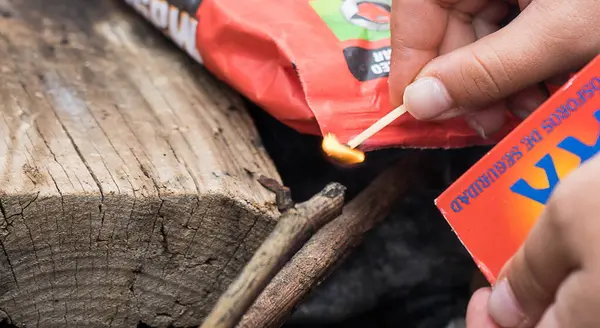 Ahşap bir kütük ve arka planda çubuklarla ateş yakan bir adam. Kibrit bir kişinin elinde..