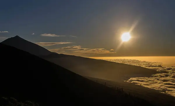 Güneşin tepenin üstünde yükseldiği sakin bir dağ manzarası bulutların üzerine sıcak bir parıltı saçıyor. Sahne bir huzur ve doğal güzellik hissi yayar..
