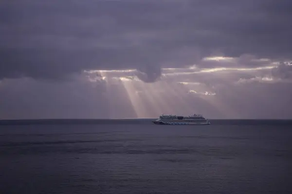 Ağır bulutlu bir gökyüzünün altında bulutları delip geçen güneş ışınlarıyla durgun sularda yol alan bir yolcu gemisinin sakin bir görüntüsü. Huzurlu deniz ve dramatik gökyüzü arasındaki zıtlık büyüleyici bir görsel deneyim yaratıyor..