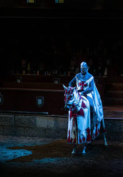 Beyaz ve kırmızı zırhlı bir şövalye, karanlık bir arenadaki spot ışıklarıyla aydınlatılmış bir atın üzerinde oturur. Sahne gizemli ve eski bir geleneği çağrıştırıyor..