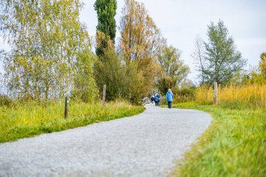 Bir adam arka planda bir göl ve ağaçlarla parkta koşuyor. Arkasında bir kadın yürüyor.