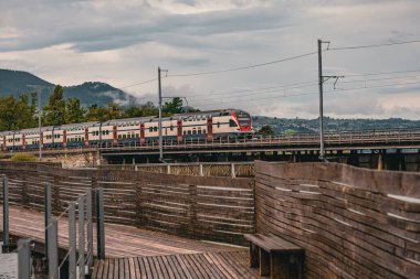 İsviçre 'nin Rapperswil kentindeki tahta yaya geçidi yakınlarındaki bir demiryolu köprüsünden geçen çift katlı modern İsviçre treni (SBB). Bulutlu bir günde, manzaralı dağ arka planında yakalanmış..