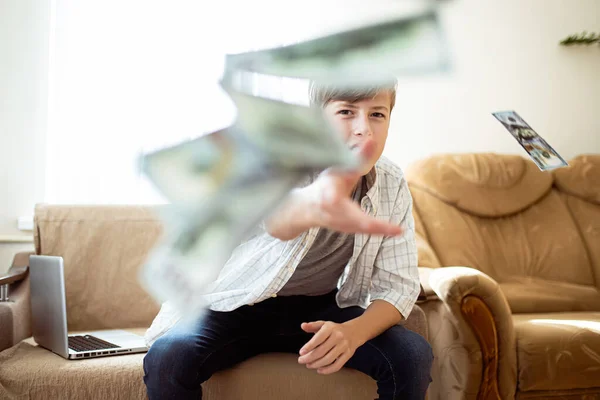 Boy flush with money. Boy throwing money around the room while sitting with laptop by the window, teenager wastes money. Boy happily scatters banknotes